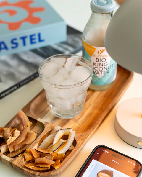 Holztablett mit Kokoschips, einem Glas mit Eis und einer Flasche Kumaio Bio King Coconut Water, daneben ein Smartphone mit geöffneter Musik-App.