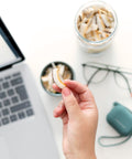 Nahaufnahme einer Hand, die eine knusprige Kokoschips-Scheibe hält, mit einem offenen Laptop und einem Glas voller Kokoschips im Hintergrund. Perfekter Snack für unterwegs oder im Büro, ideal für gesundheitsbewusste Genießer.